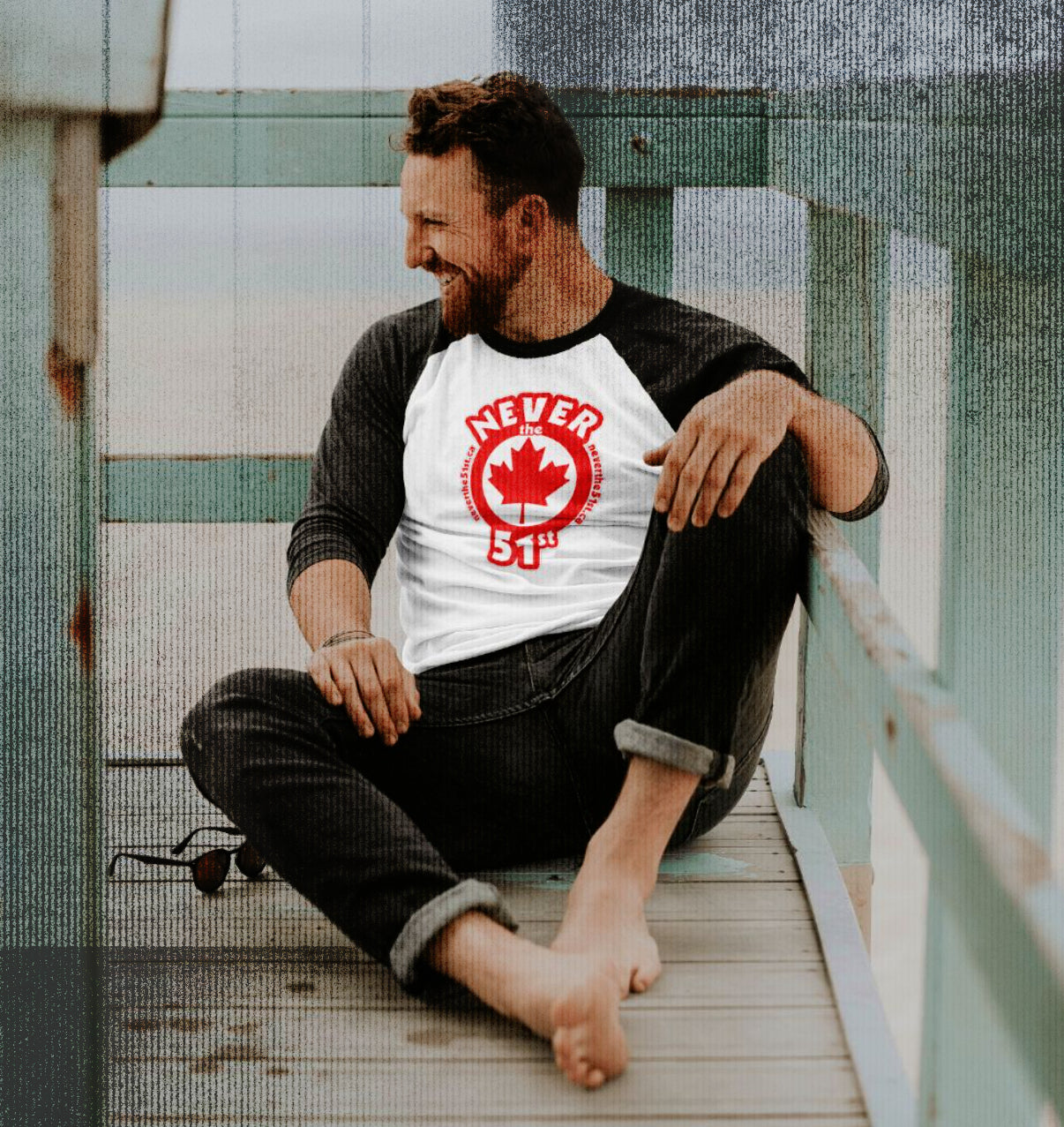 Man wearing a white and black Never the 51st 3/4 sleeve tee with red maple leaf logo, sitting barefoot on a beachside deck and smiling