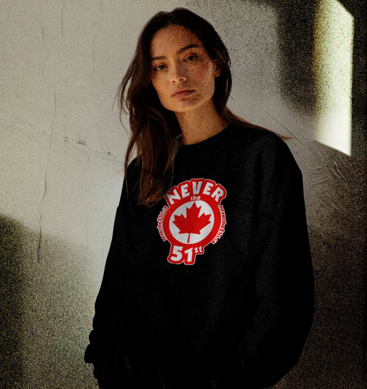 Woman wearing a black Never the 51st crewneck sweater with red maple leaf logo, standing in natural light against a neutral wall
